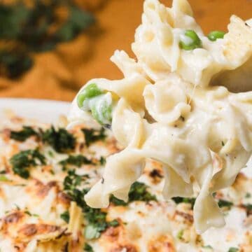 A fork lifts creamy pasta bake with peas and cheese from a casserole dish, garnished with herbs.