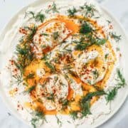 Whipped yogurt topped with poached eggs, paprika oil, and fresh dill on a white plate.
