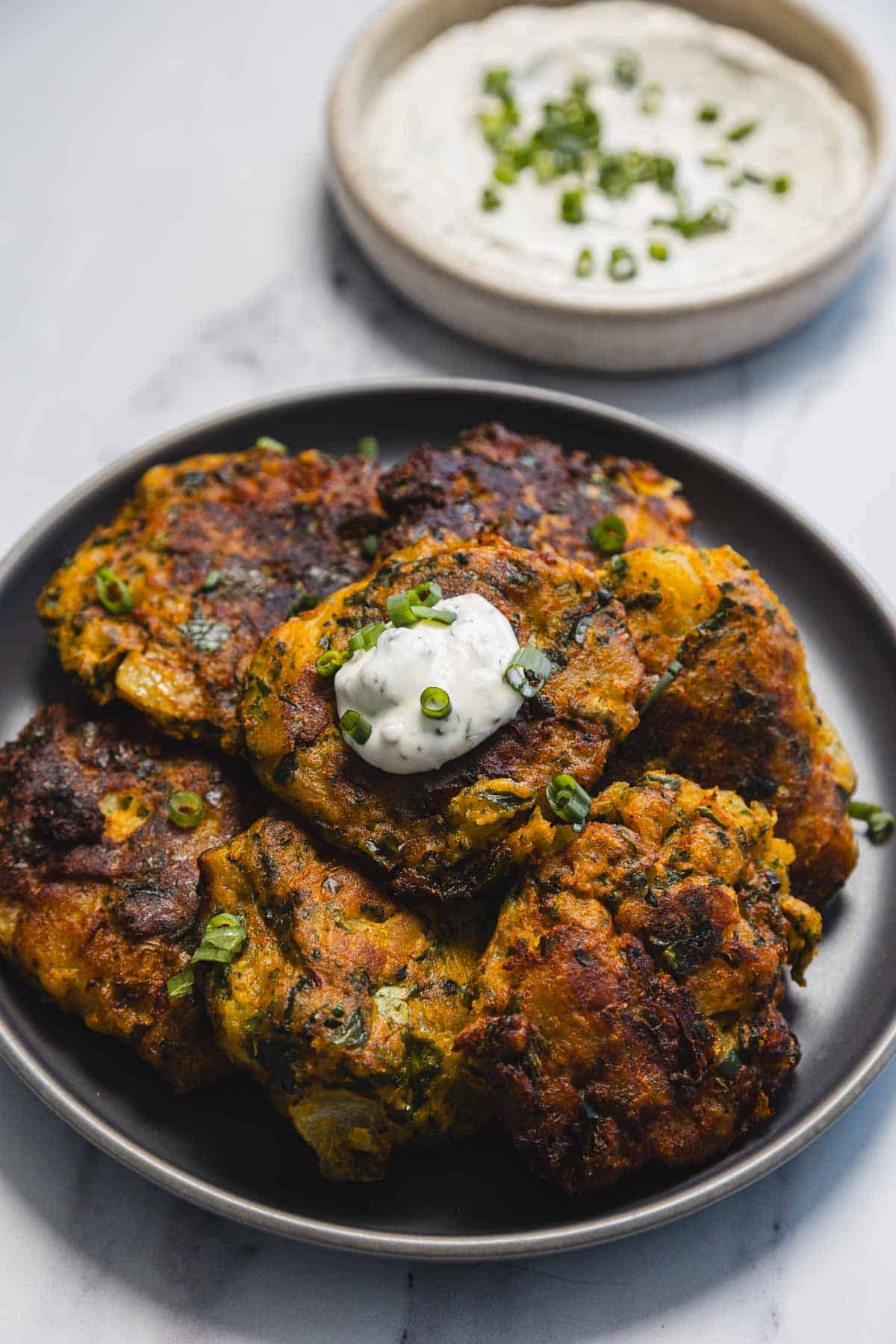 A plate of browned Aruk&mdash;Iraqi Style Latkes vegetable fritters topped with a dollop of white sauce and chopped green onions, with a bowl of dipping sauce garnished with green onions in the background.