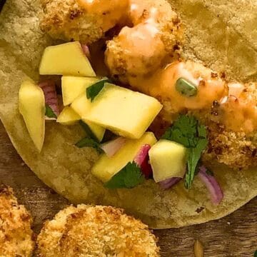 A taco with breaded shrimp, diced mango, red onion, cilantro, and orange sauce on a corn tortilla.