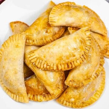 A white plate filled with golden brown, baked empanadas on a wooden table with flowers nearby.