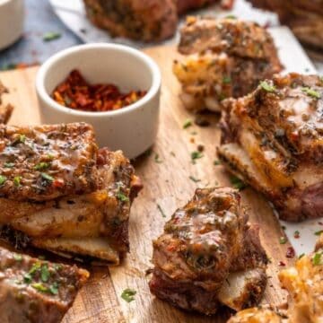 Seasoned and cooked beef short ribs on a wooden board, garnished with herbs, with sauce and spices nearby.