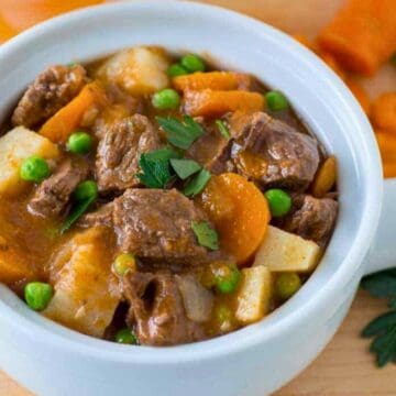 A bowl of beef stew with carrots, peas, potatoes, and parsley on a wooden table.