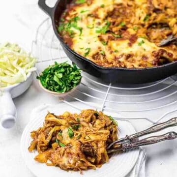 A plate of cheesy baked pasta with herbs, next to a skillet of pasta, shredded cheese, and chopped parsley.