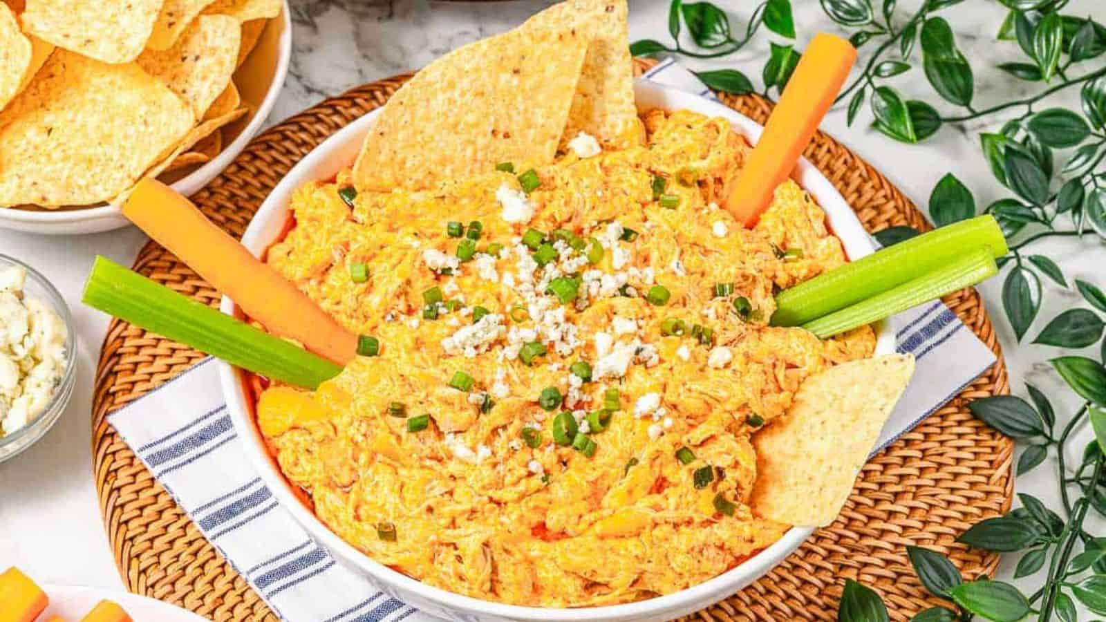 Bowl of buffalo chicken dip topped with green onions, surrounded by celery, carrot sticks, and tortilla chips&mdash;perfect for party dips and snacking.
