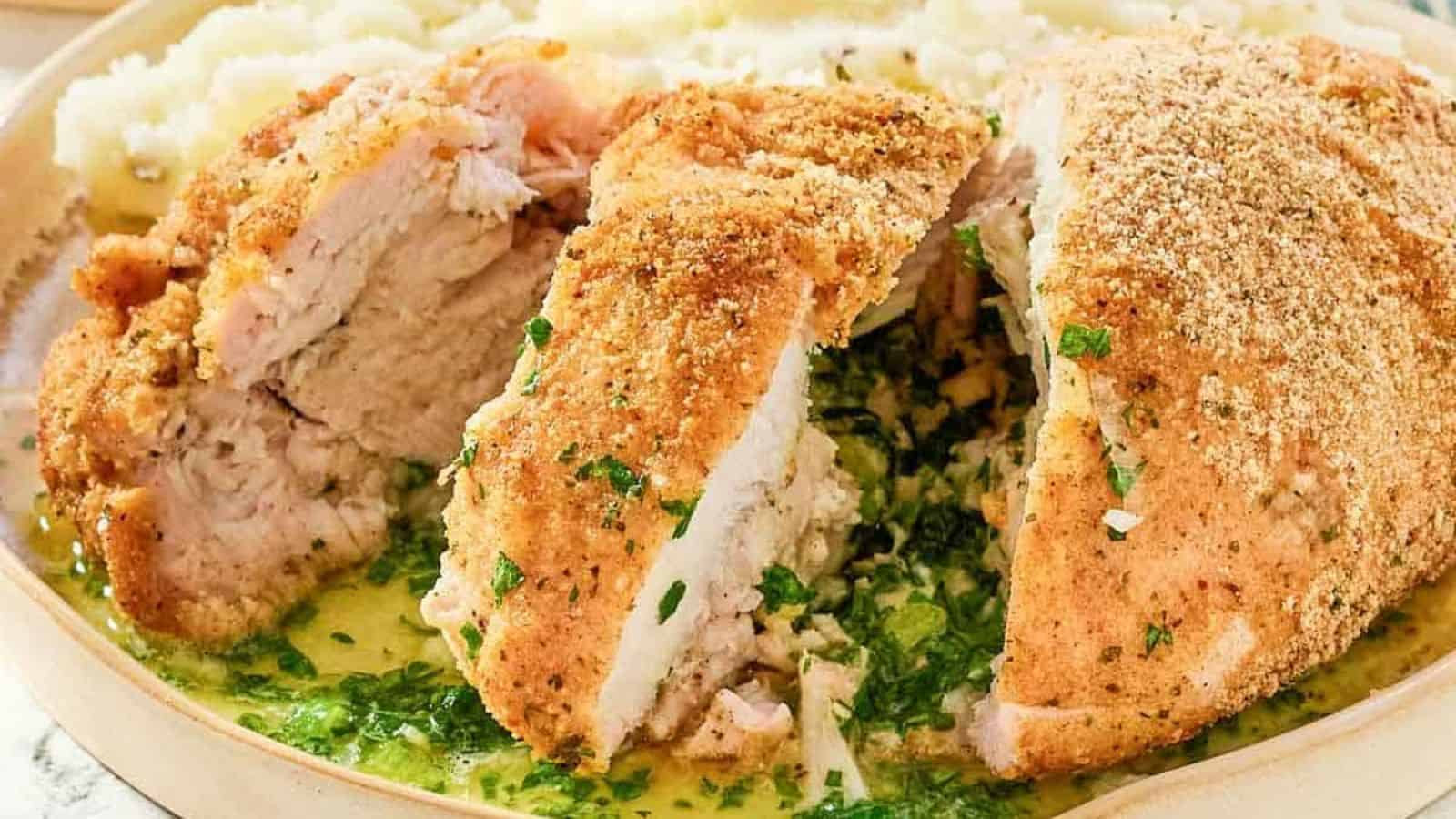 Breaded chicken breast sliced open, revealing a herbed buttery filling, served with mashed potatoes.