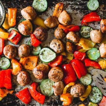 Roasted sausage, zucchini, and colorful bell peppers come together on a baking sheet with dried herbs in a small dish—perfect for fans of easy one-pan recipes.