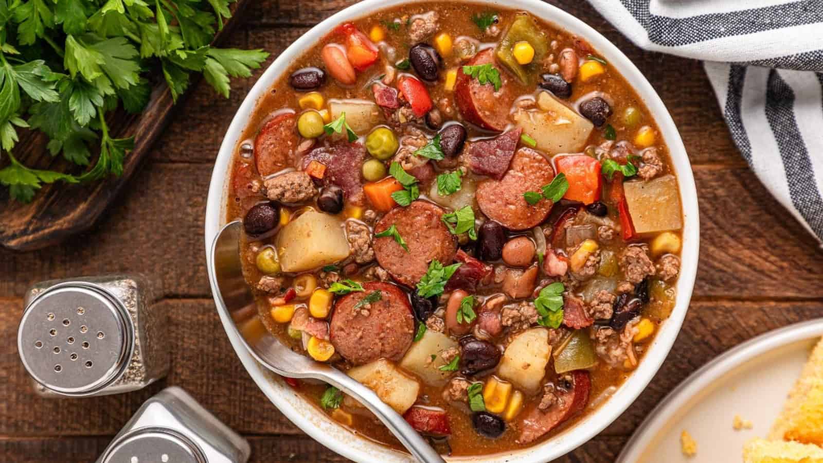 A pot of mixed vegetable and sausage stew with corn, beans, potatoes, and herbs, accompanied by sliced cornbread on the side.