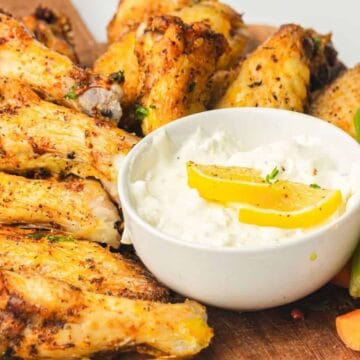 A pile of seasoned chicken wings with dip, lemon slices, and carrot and celery sticks on a wooden board—perfect for casual chicken dinners.