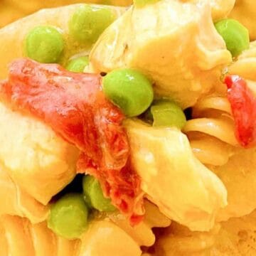 Close-up of a wooden spoon holding pasta with peas, chicken pieces, and red bell pepper strips.