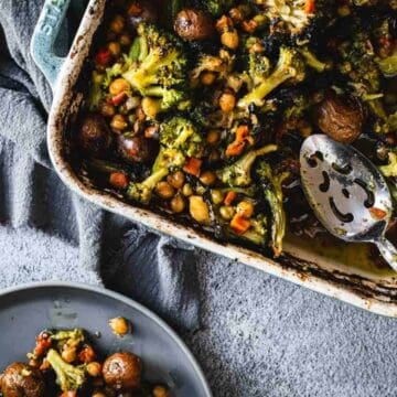A baking dish of roasted broccoli, mushrooms, and chickpeas next to a plate with a serving and a serving spoon.