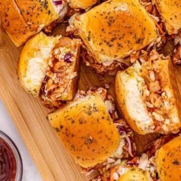 BBQ chicken sliders with coleslaw on a wooden board, served with chips and a bowl of barbecue sauce.