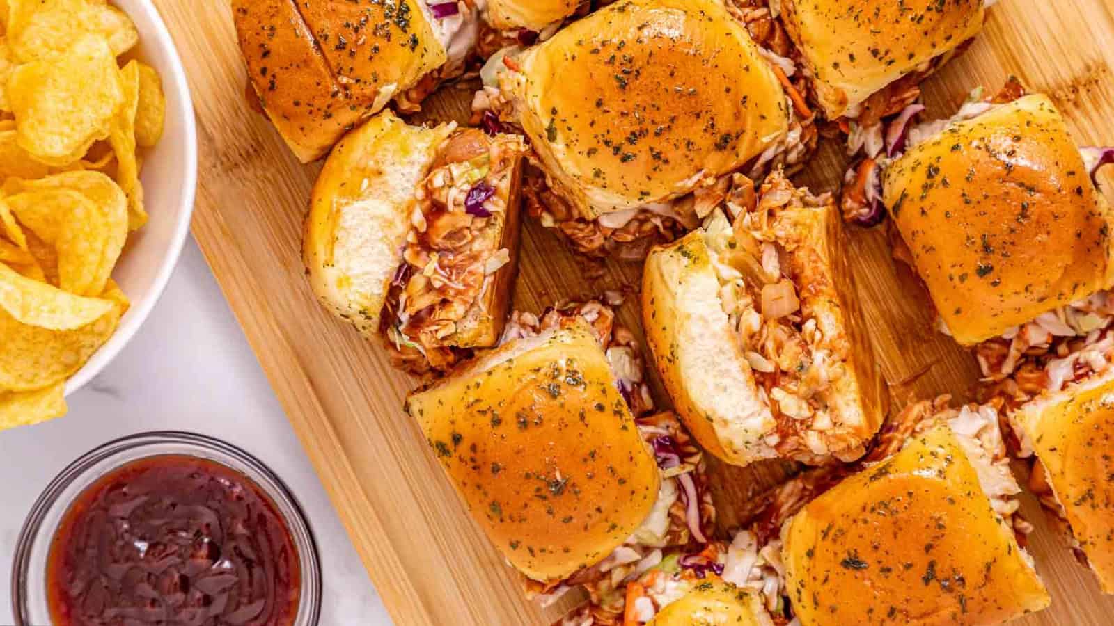 BBQ chicken sliders with coleslaw on a wooden board, served with chips and a bowl of barbecue sauce.