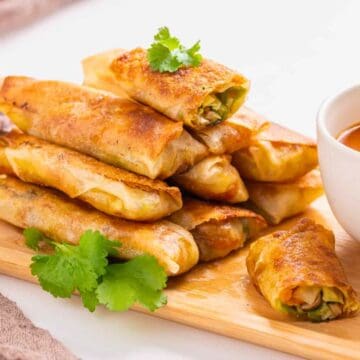 A stack of crispy spring rolls on a wooden board with dipping sauce and cilantro garnish.