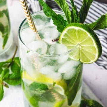 A glass of mojito with lime, mint leaves, ice, and a straw on a table with a striped cloth nearby.
