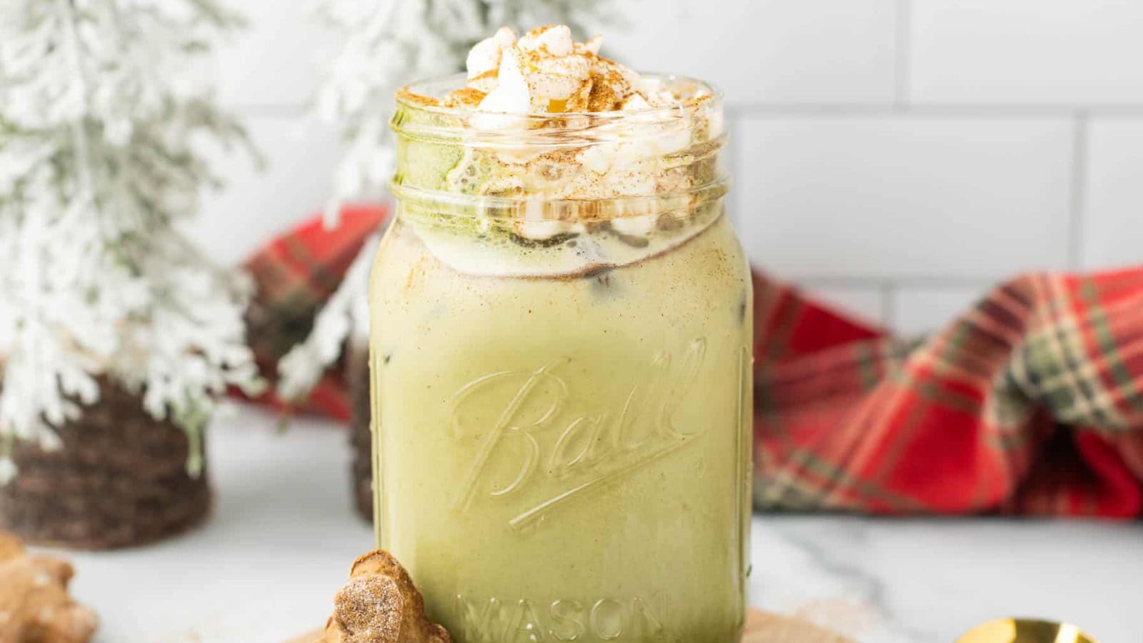 A mason jar of iced matcha latte topped with whipped cream and cinnamon, with holiday decor in the background.