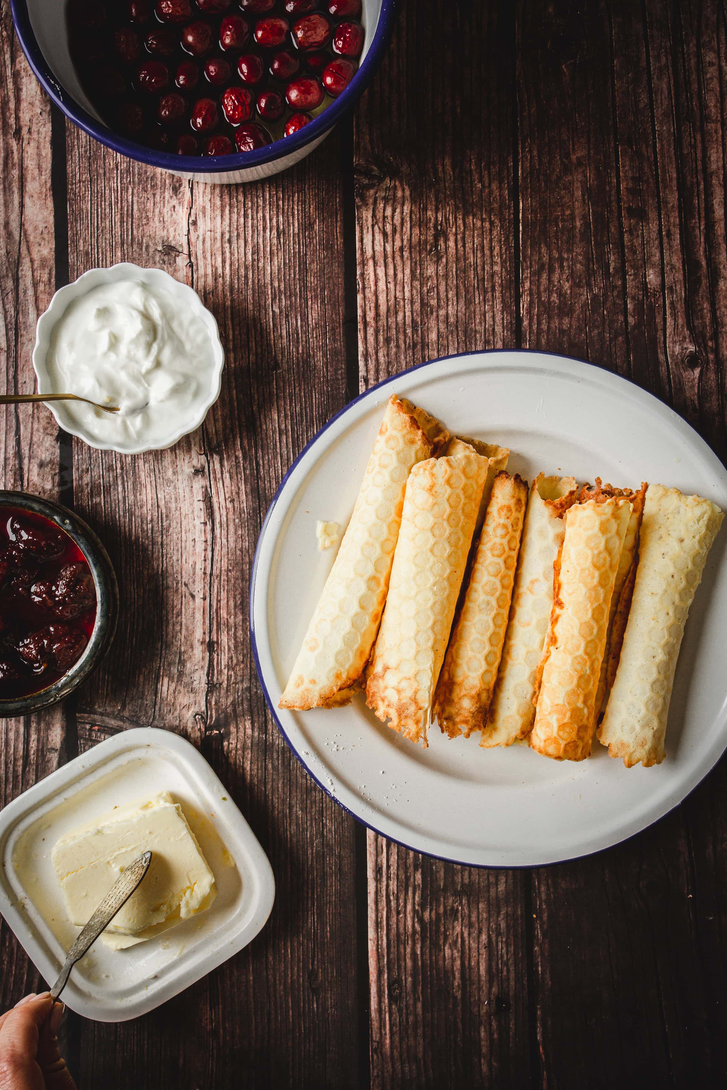 Rolled crepes on a plate evoke the charm of traditional Russian rolled wafers, served with bowls of cherries, jam, sour cream, and a dish of butter on a wooden table.