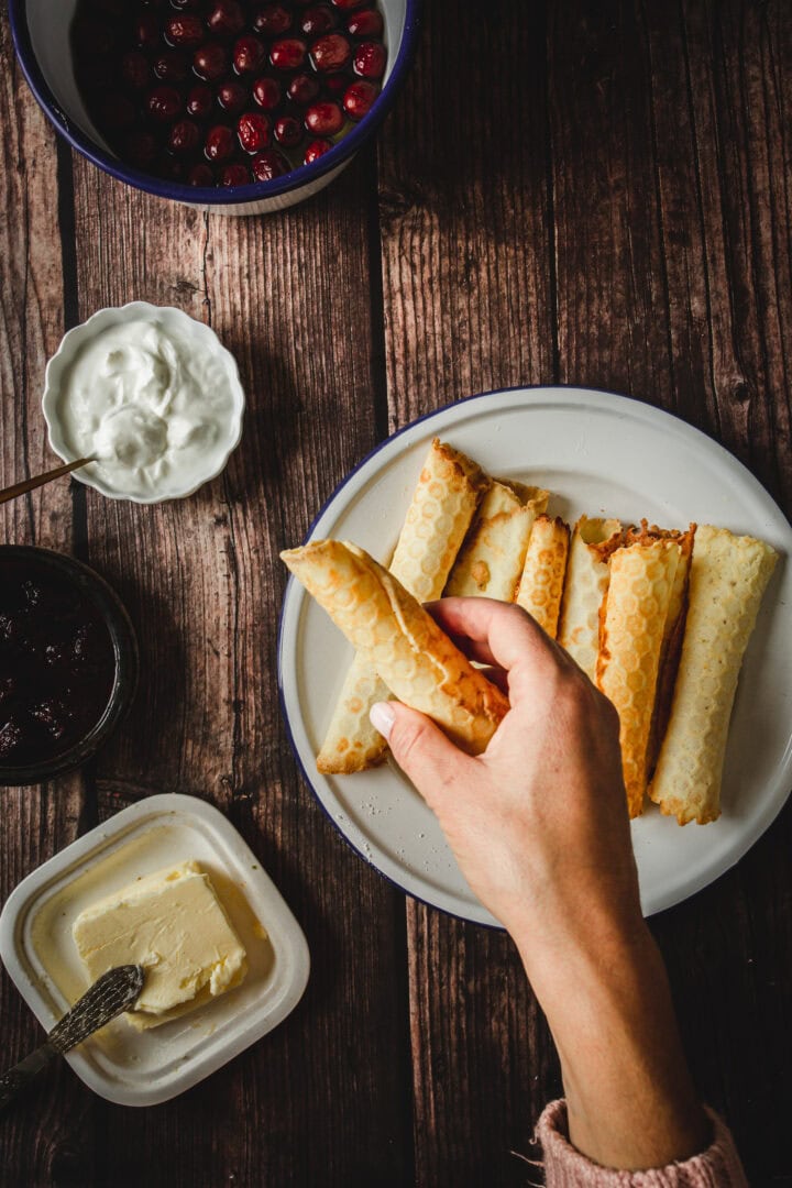 A hand holds a rolled crepe over a plate of crepes, with butter, jam, yogurt, and berries nearby-reminiscent of traditional Russian rolled wafers.