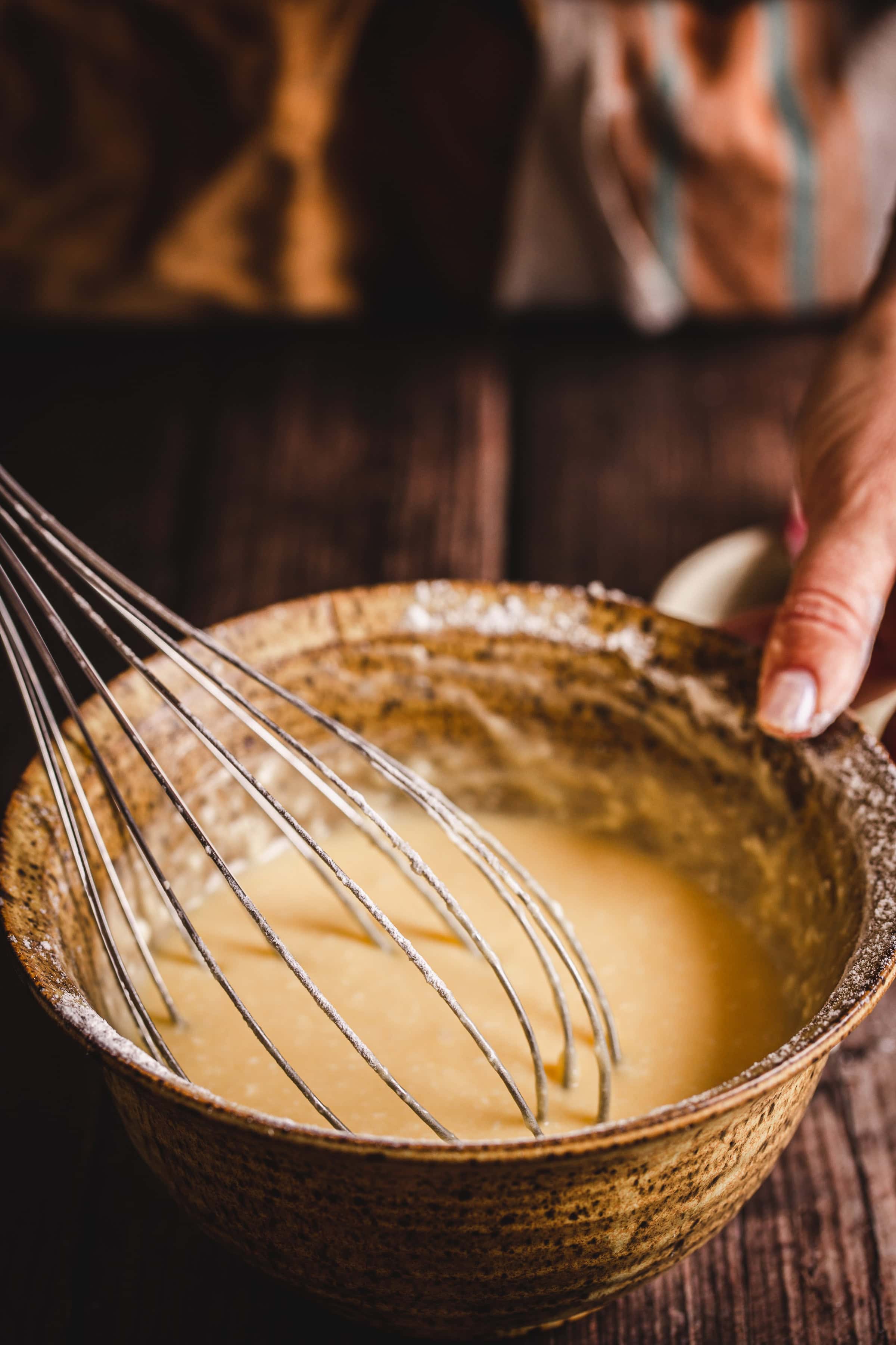 A hand holds a whisk stirring batter for traditional Russian rolled wafers in a rustic brown bowl on a wooden surface.
