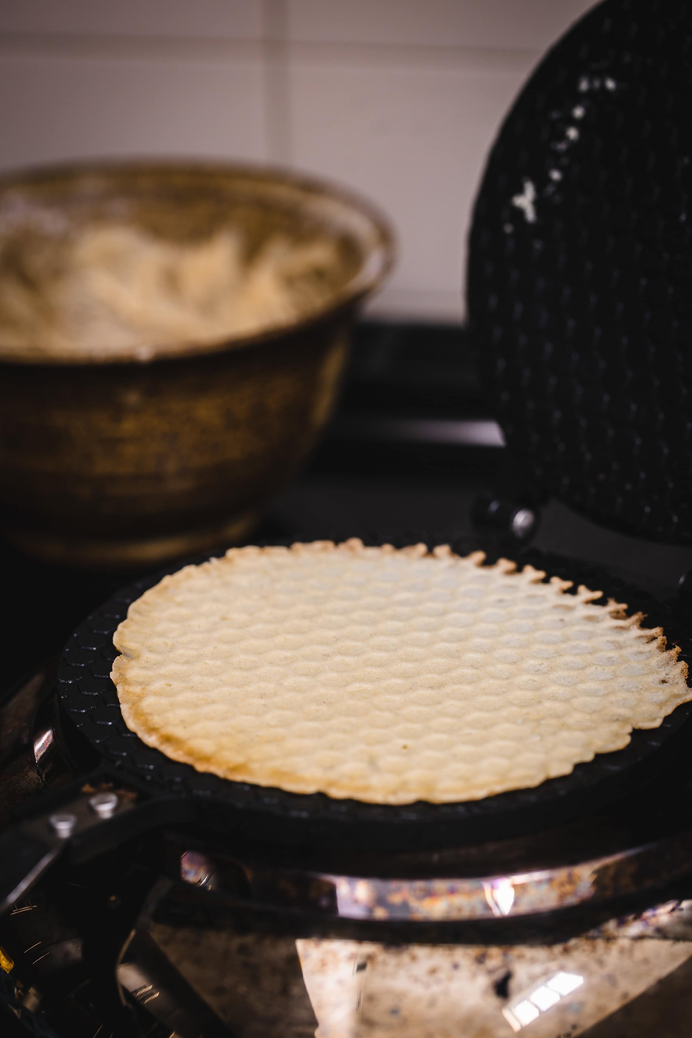 A thin waffle, perfect for making traditional Russian rolled wafers, cooks on a waffle iron, with a bowl of batter in the background.