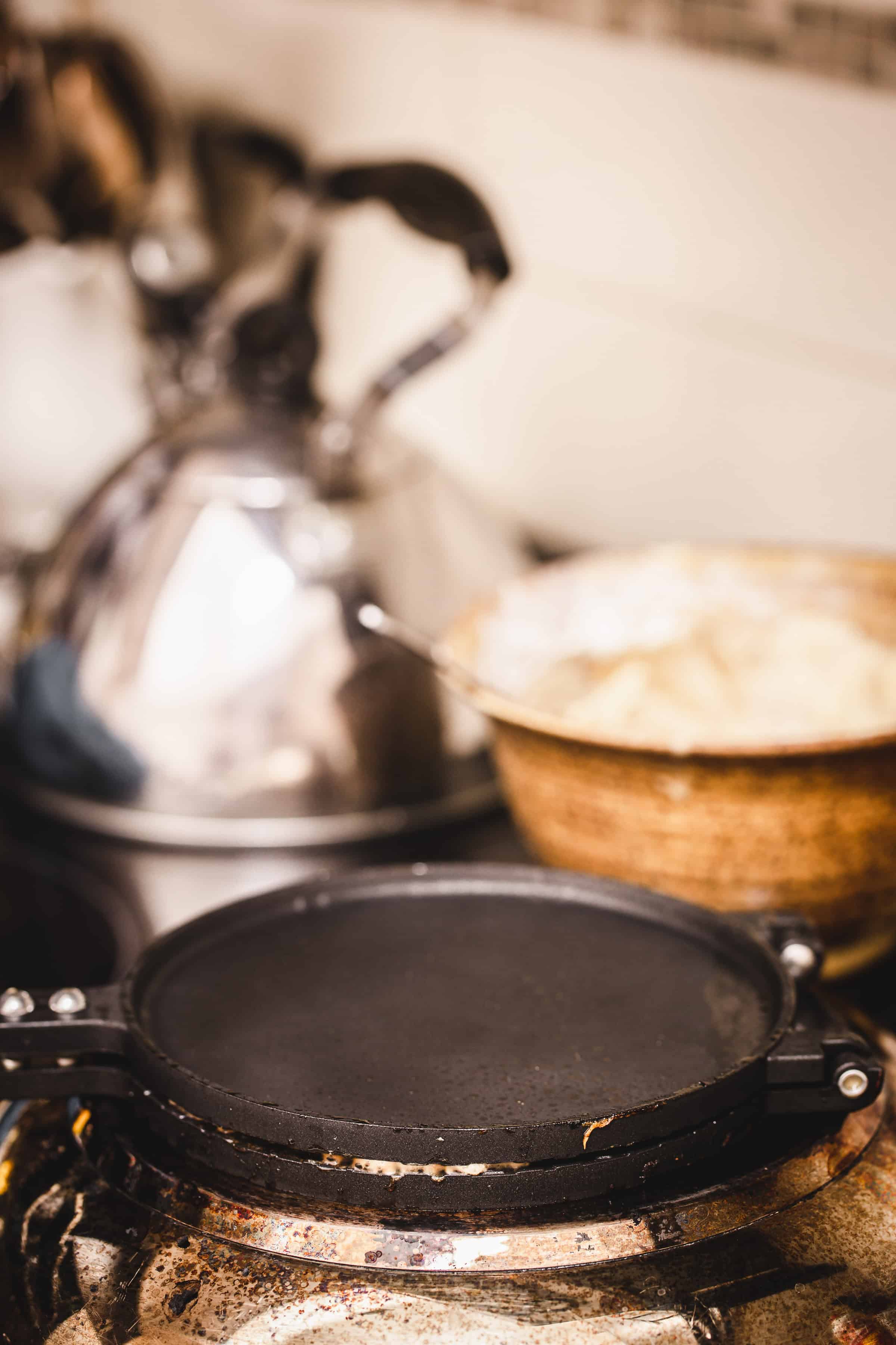 A black griddle on a stovetop, ideal for making traditional Russian rolled wafers, with a metal kettle and a mixing bowl in the background.