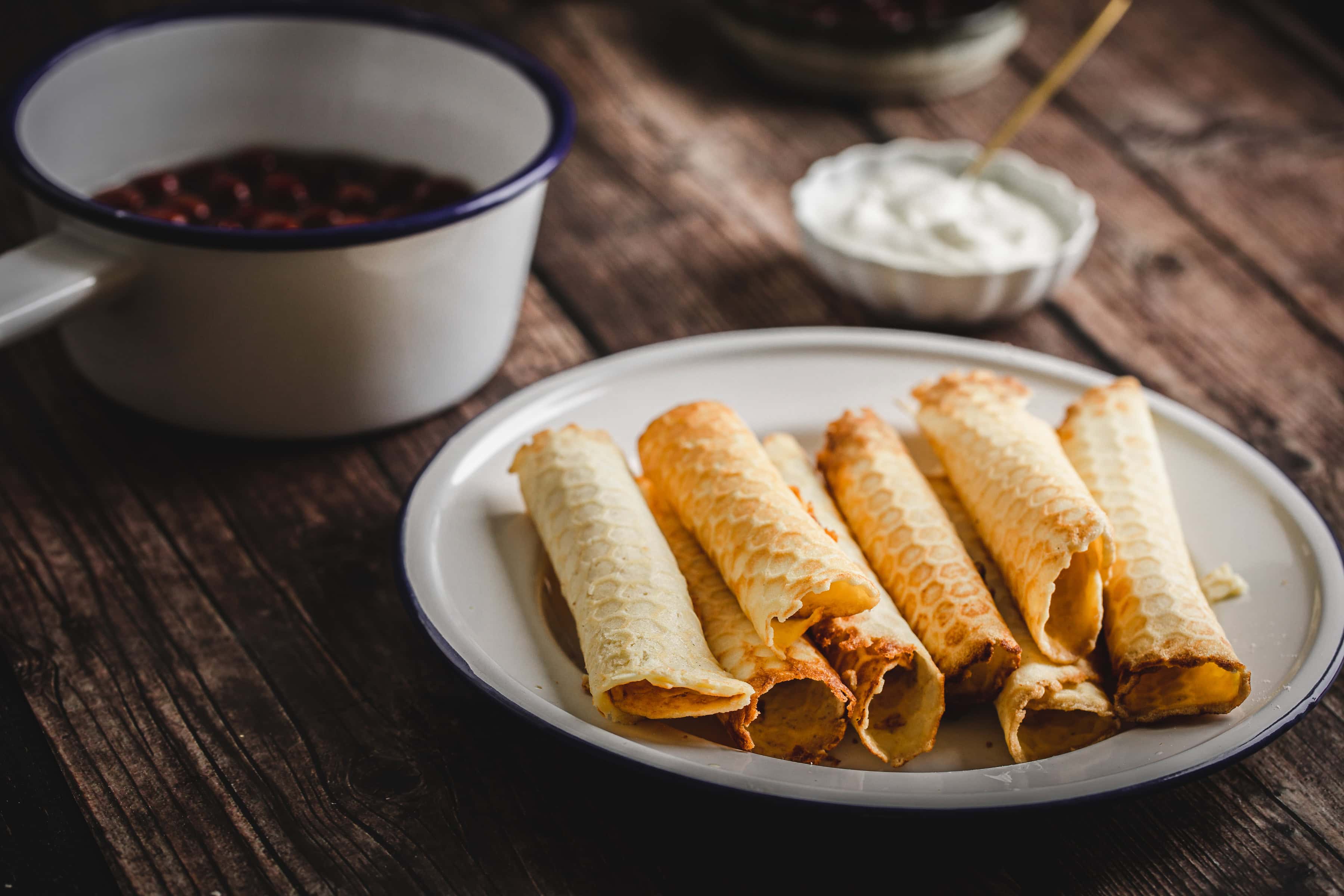 Traditional Russian rolled wafers on a plate, served with a pot of red sauce and a bowl of cream atop a rustic wooden table.