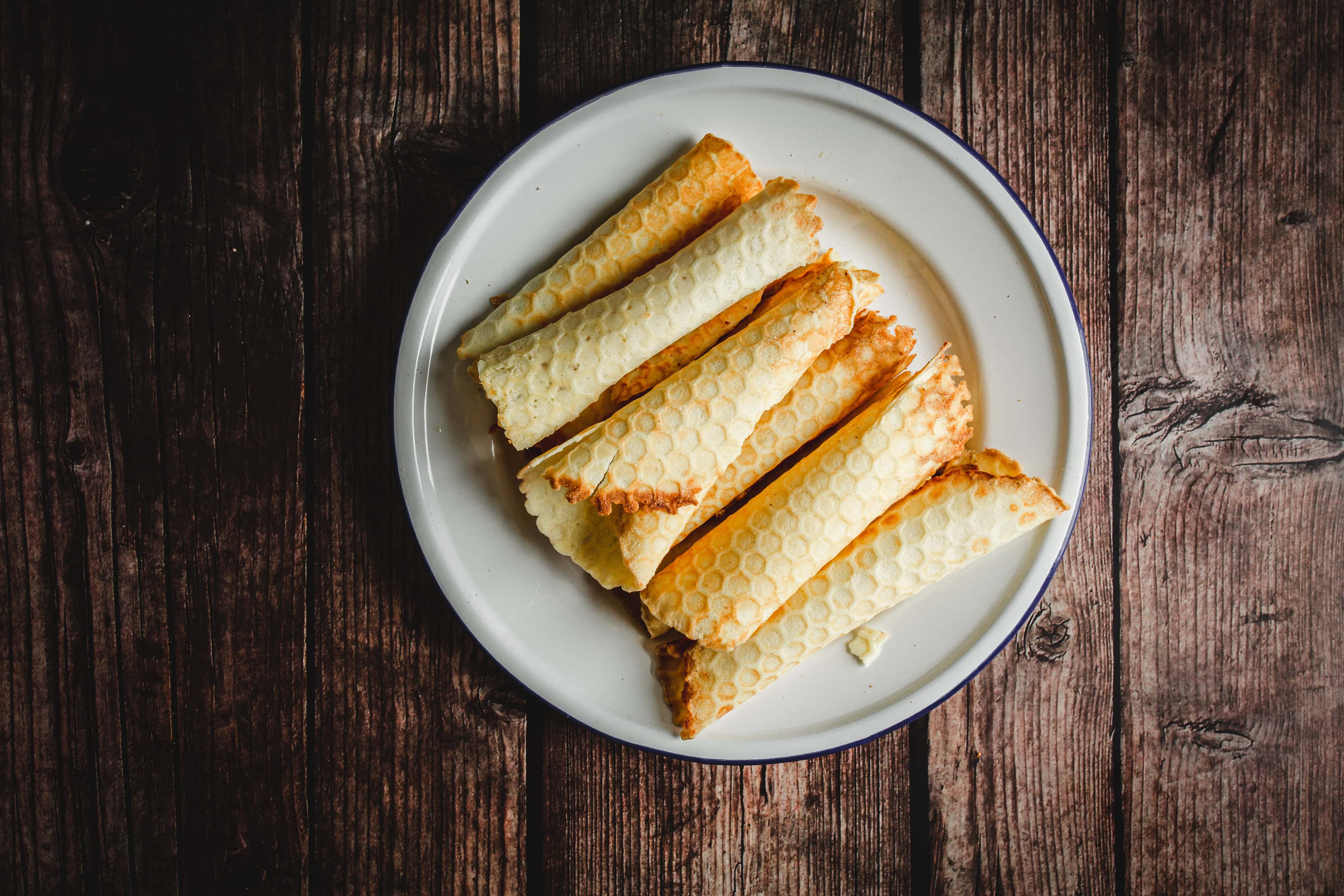 Six traditional Russian rolled wafers with a honeycomb pattern are neatly arranged on a white plate atop a dark wooden surface.