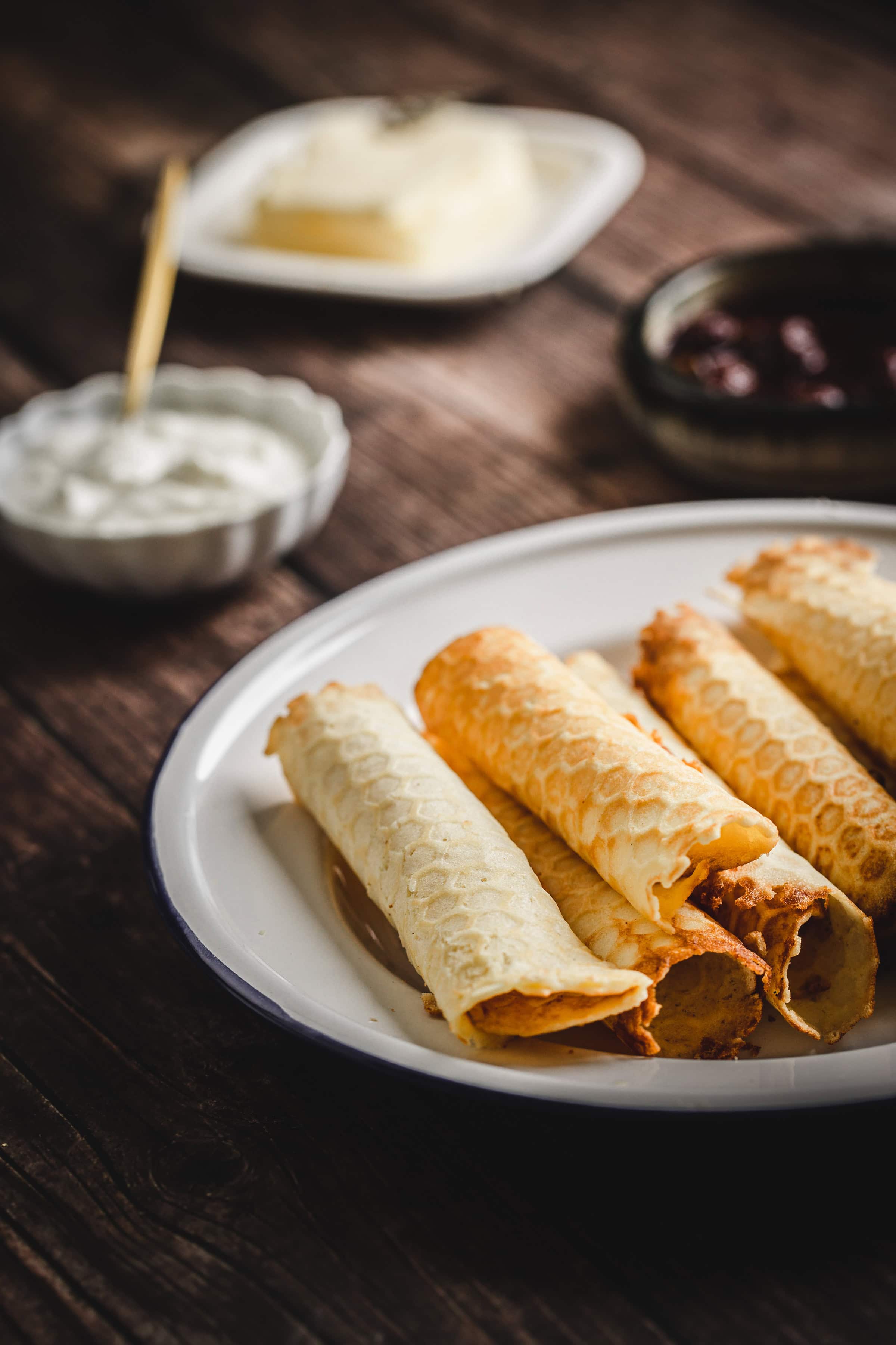 Rolled waffles on a white plate, reminiscent of traditional Russian rolled wafers, served with small bowls of cream, butter, and jam in the background.