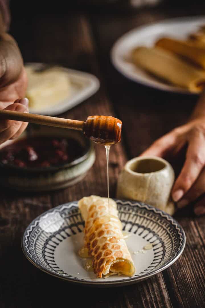 A hand drizzles honey onto traditional Russian rolled wafers on a patterned plate, with more pancakes in the background.