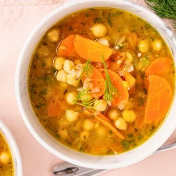 A bowl of chickpea and vegetable soup with carrots, herbs, and a spoon on a pink cloth.