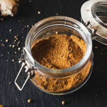A glass jar of ground spice with an open lid, ginger root, and a spoon on a dark surface.