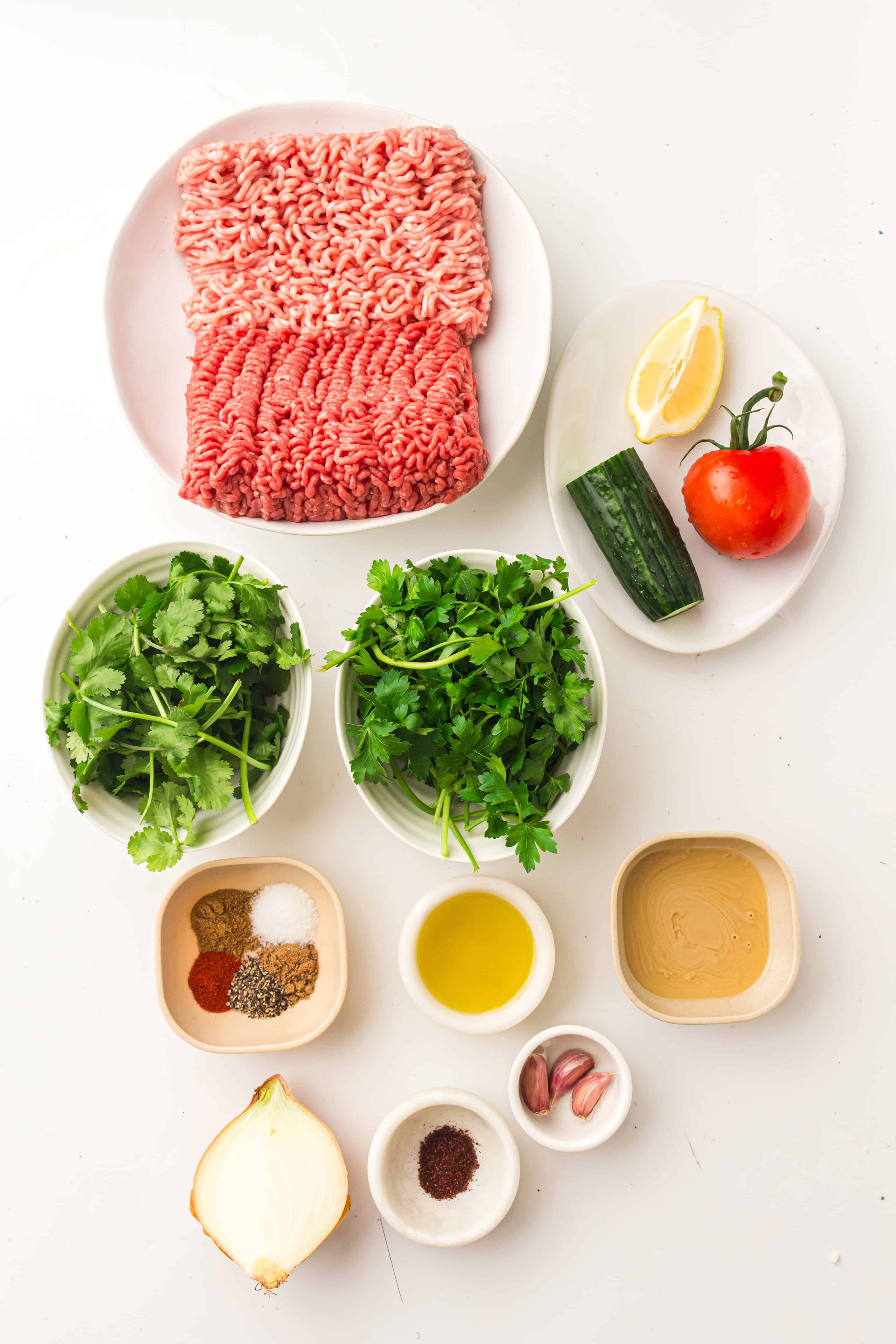 Ingredients for a recipe, including ground meat, herbs, spices, onion, garlic, cucumber, tomato, lemon, and oil.