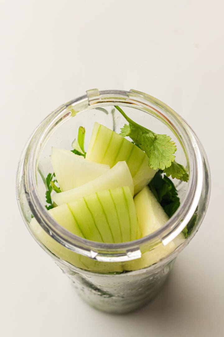 Chopped onions, cilantro, and cucumber in a blender cup, ready to be blended, on a white background.