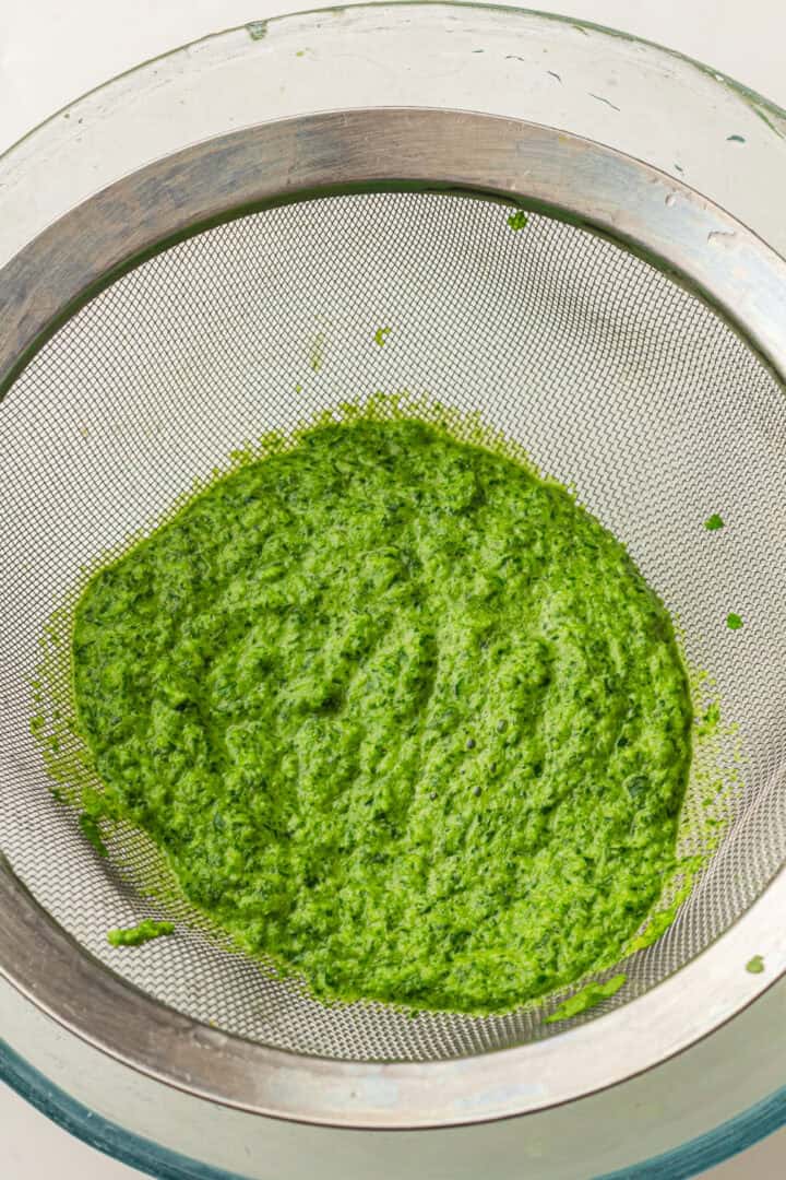 Bright green blended herb mixture sits in a fine mesh strainer over a glass bowl.
