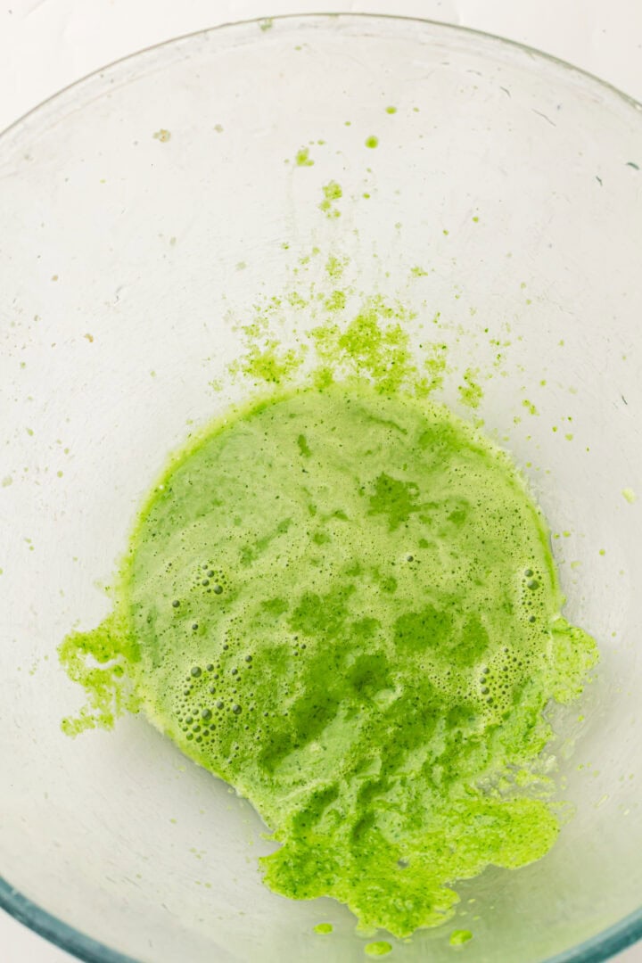 Bright green liquid and foam in a clear glass bowl, viewed from above on a white background.
