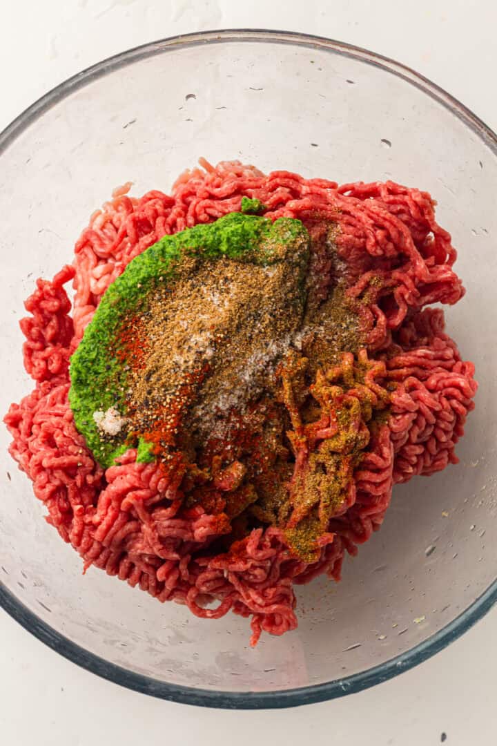 Raw ground meat in a glass bowl topped with green paste and assorted spices, ready to be mixed.