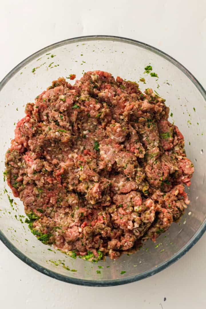 Ground meat mixed with herbs and spices in a clear glass bowl on a white surface.