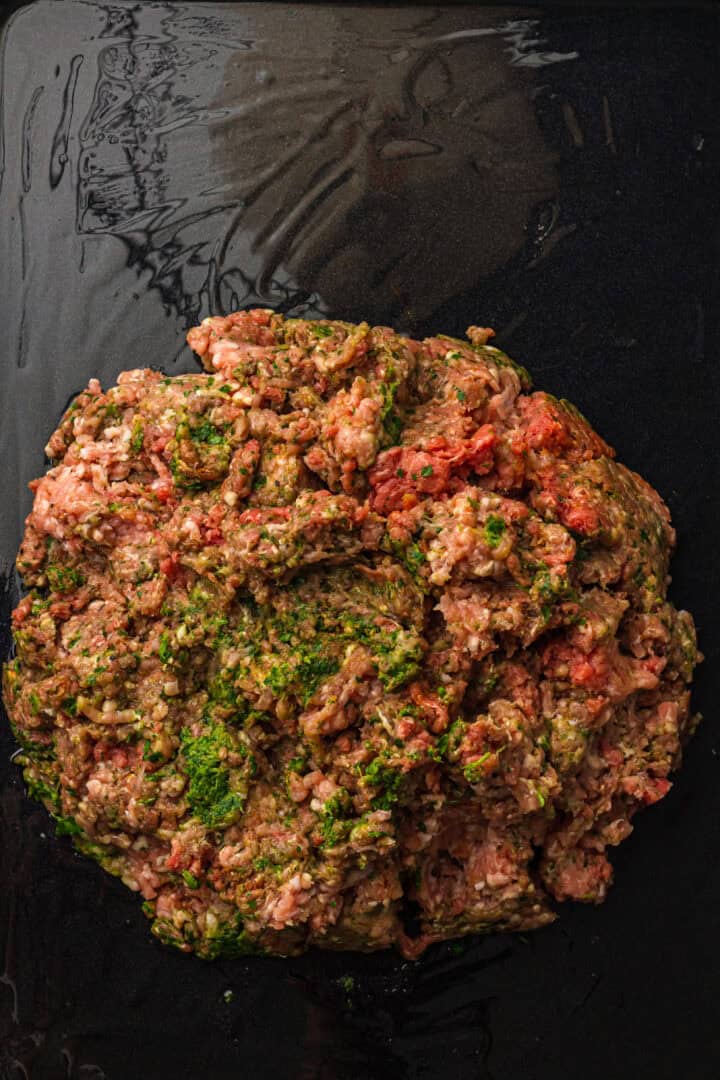 A pile of seasoned raw ground meat with herbs on a dark, oily surface.