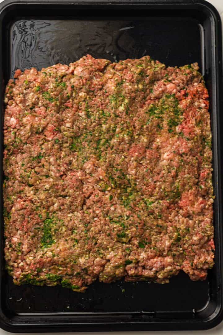 Raw seasoned ground meat spread evenly on a black baking tray, ready to be cooked.