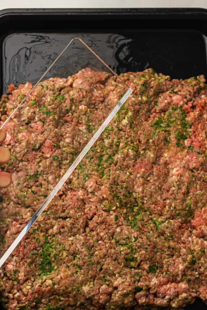 A hand holds a clear rectangular tool over seasoned ground meat spread in a black baking tray.
