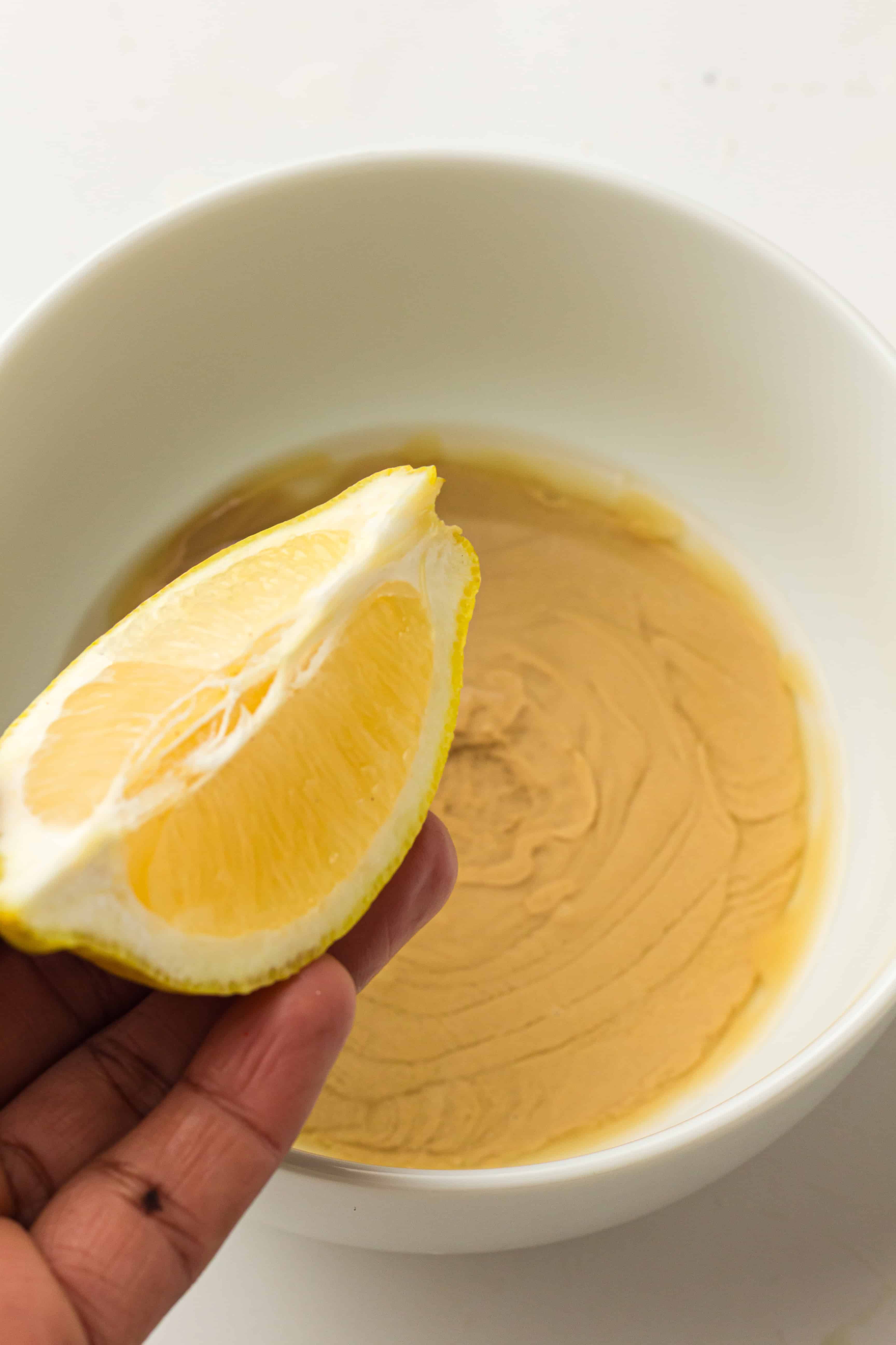 Hand holding a lemon wedge over a white bowl filled with creamy light brown sauce.