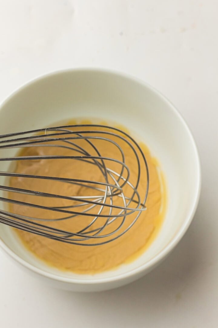 A metal whisk rests in a white bowl filled with a creamy brown mixture.