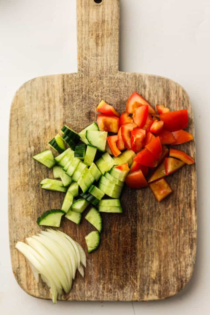 Chopped cucumber, tomato, and onion on a wooden cutting board against a light background.