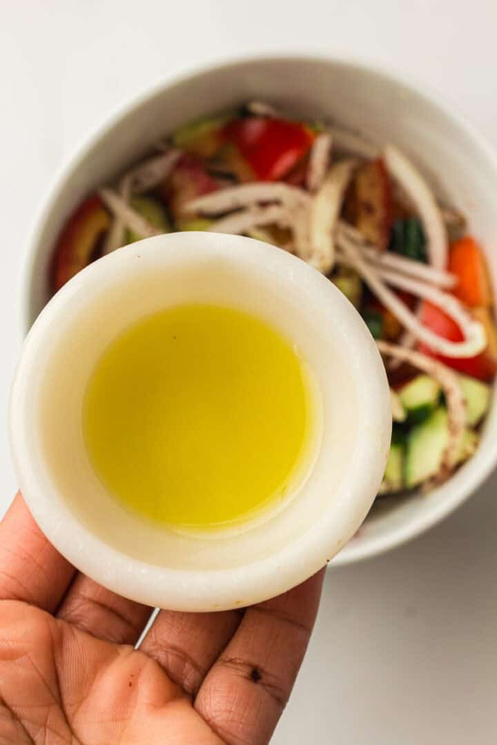A hand holds a small white bowl of olive oil above a bowl of chopped salad vegetables.