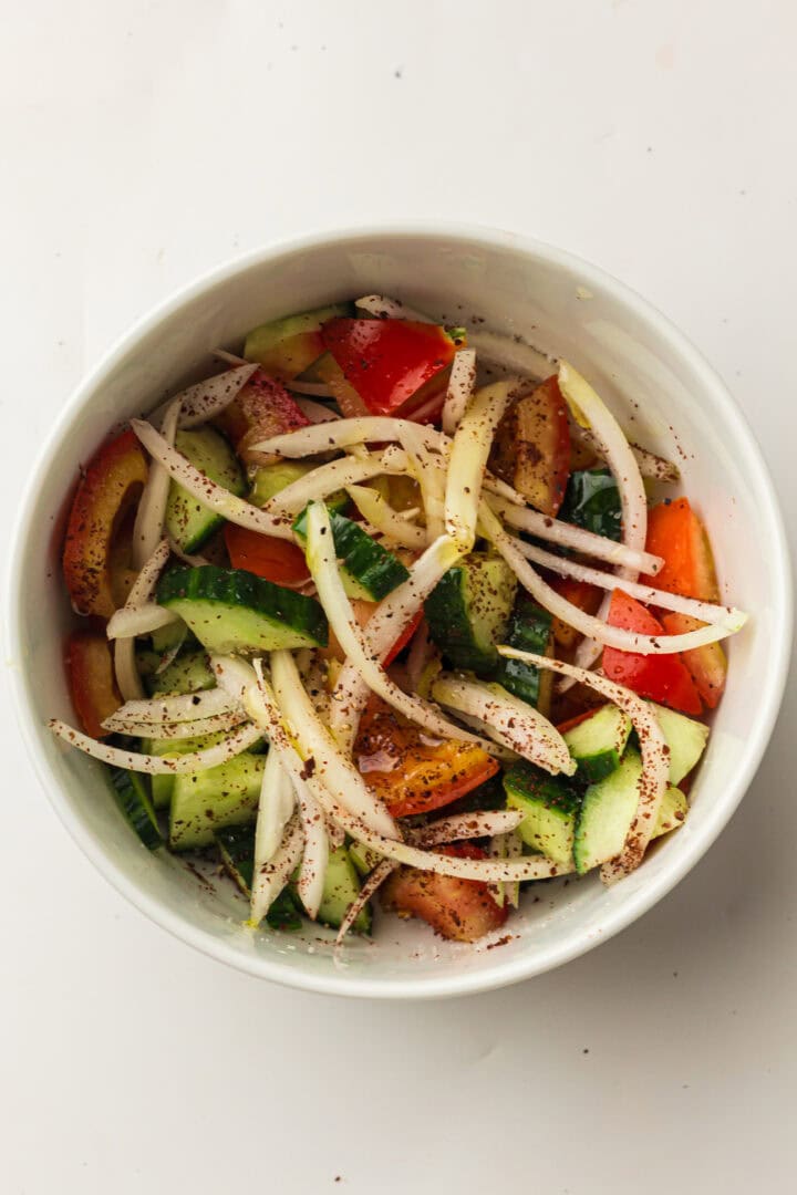 A bowl of salad with sliced onions, cucumbers, and tomatoes, sprinkled with seasoning.