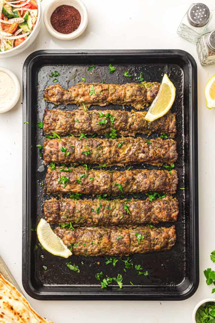 Six grilled kebabs garnished with parsley and lemon wedges on a black tray, with sides and seasonings nearby.