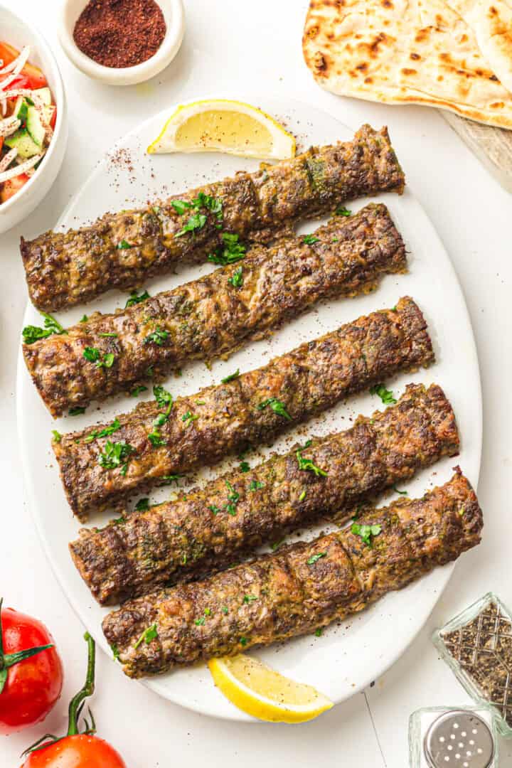 Five grilled kebabs on a white plate, garnished with herbs and lemon wedges, served with flatbread and salad.