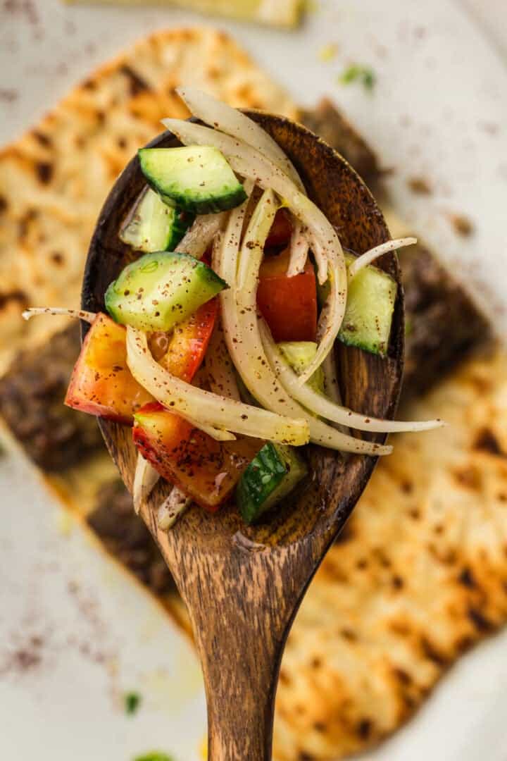 A wooden spoon holds a salad of onion, tomato, and cucumber over flatbread on a plate.