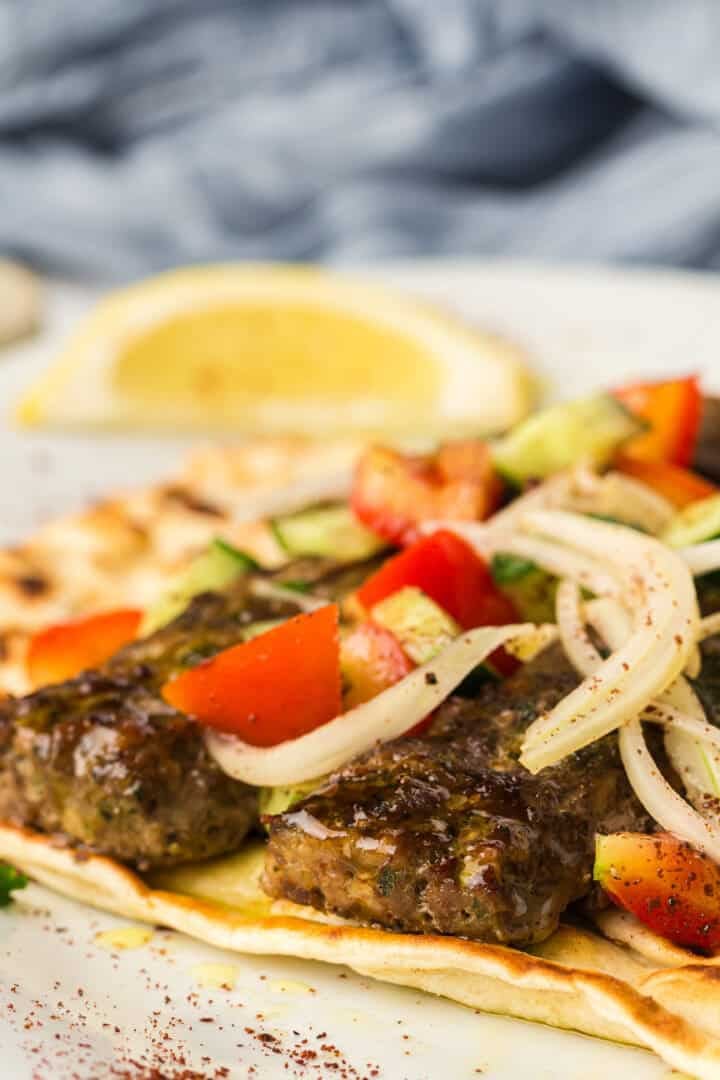 Close-up of grilled kebab on flatbread, topped with sliced onions, diced tomatoes, and a lemon wedge in the background.