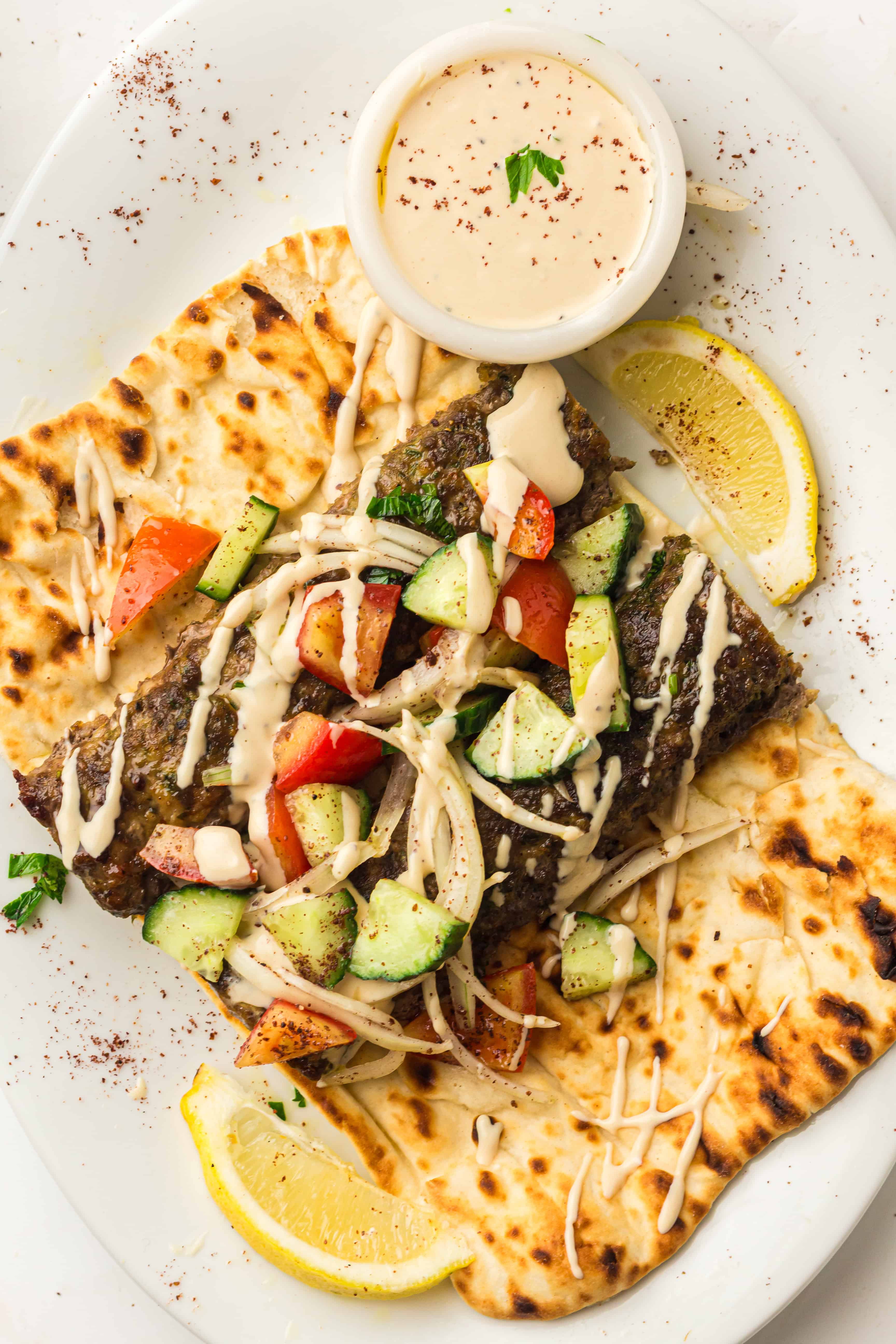 Grilled kebab on flatbread with chopped salad, tahini sauce, and lemon wedges on a white plate.
